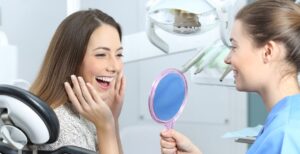 A happy woman looking in the mirror at her dental implants results, the dentist is holding the mirror.